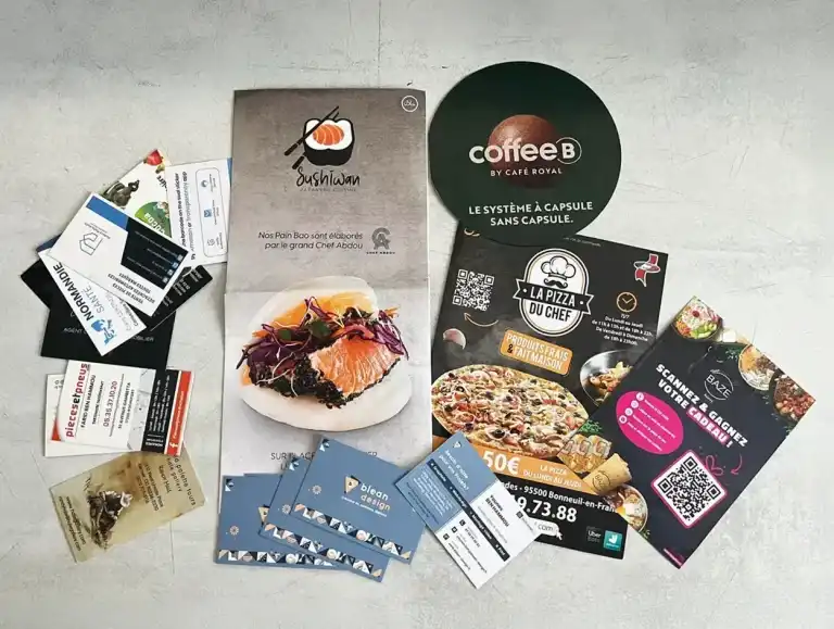 Print blean design Exemples de cartes de visite et flyers publicitaires imprimés par Blean Design : menus de restaurants, cartes de visite professionnelles, flyers promotionnels et supports marketing variés disposés sur une table claire.